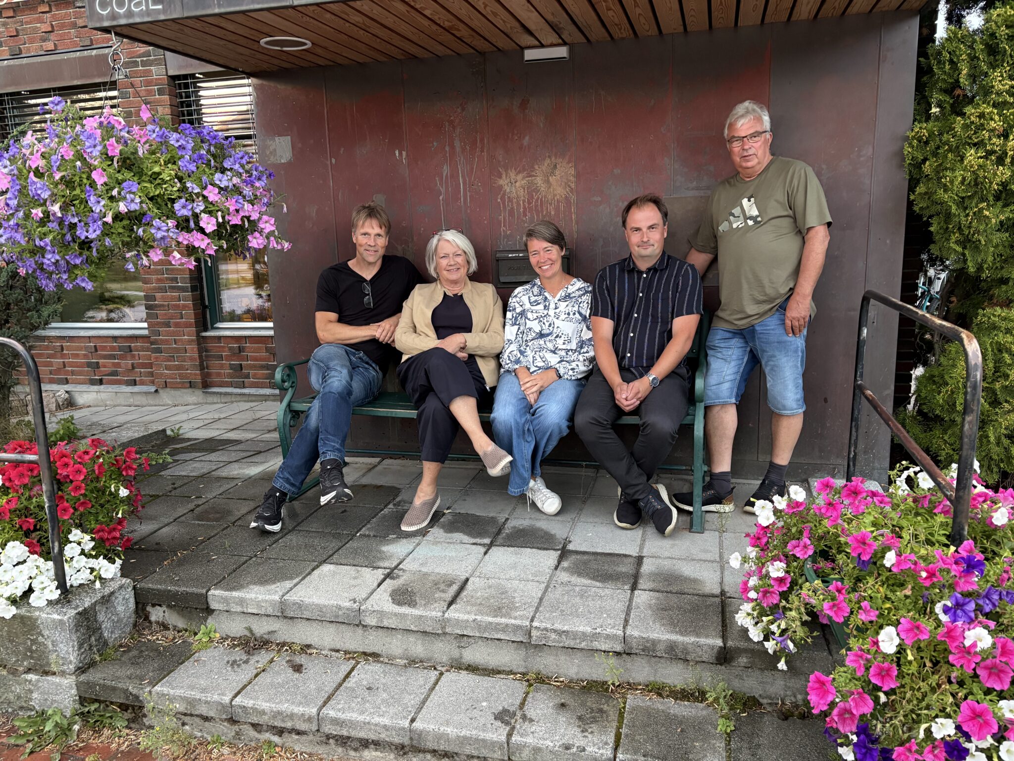 Five adults posing on a green bench outdoors beside a brick building with hanging purple and pink flowers nearby.